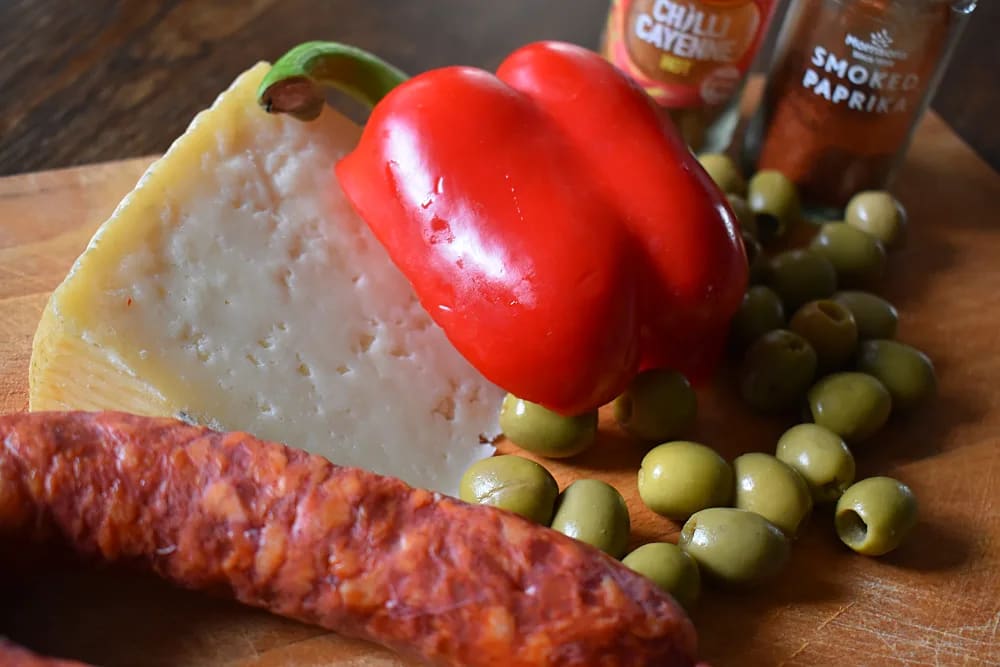 Ingredients for Olive and Chorizo Scones with Manchego Cheese laid out on a wooden board - Chorizo, half a red pepper, wedge of manchego cheese, green olives and cayenne and paprika jars.