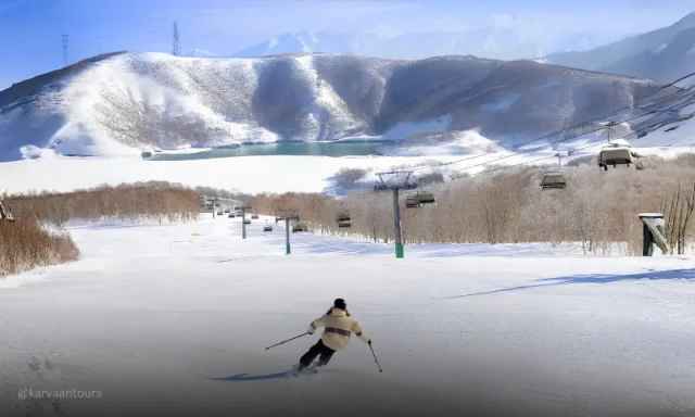 A lone skier going down a white slope toward a bright turquoise lake surrounded by snowy mountains.