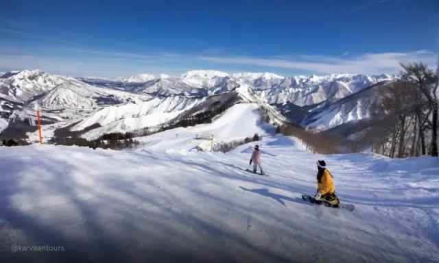 Snowboarders resting on a wide, sunny mountain ridge looking out over a massive snowy range.
