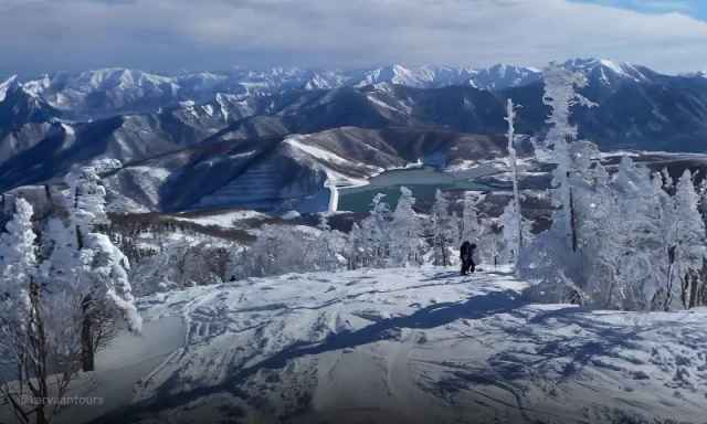 Frost-covered trees on a snowy peak overlooking a deep green lake in the valley below.