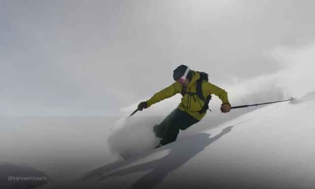 A skier in a yellow jacket aggressively kicking up a cloud of white snow on a misty slope.