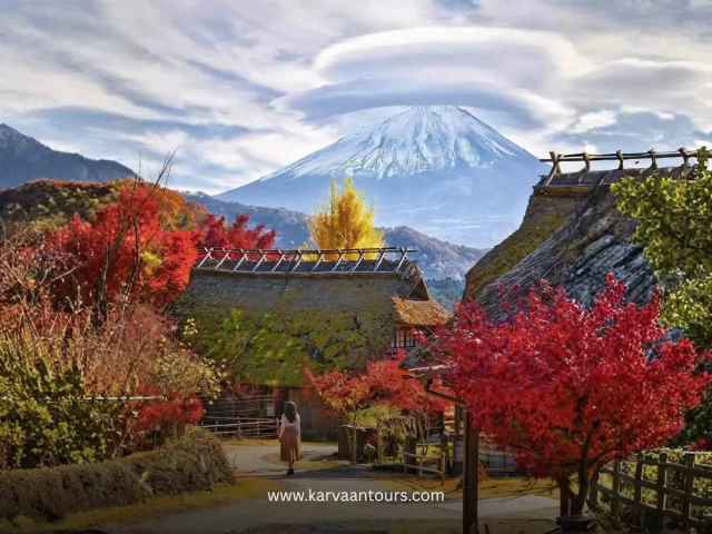A woman walking along a path in a traditional Japanese village during autumn, surrounded by vibrant red and yellow foliage, with a magnificent snow-capped Mount Fuji under a distinctive lenticular cloud in the background.