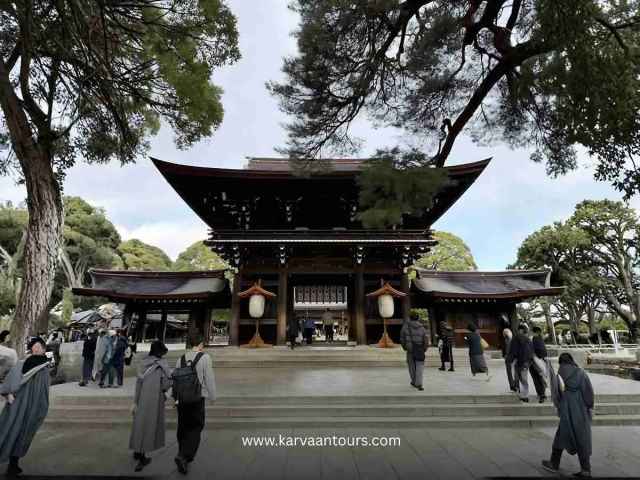 A view of the impressive main hall of Meiji Jingu Shrine in Tokyo, featuring traditional wooden architecture, large white hanging lanterns, and visitors walking through the courtyard under the shade of mature trees.
