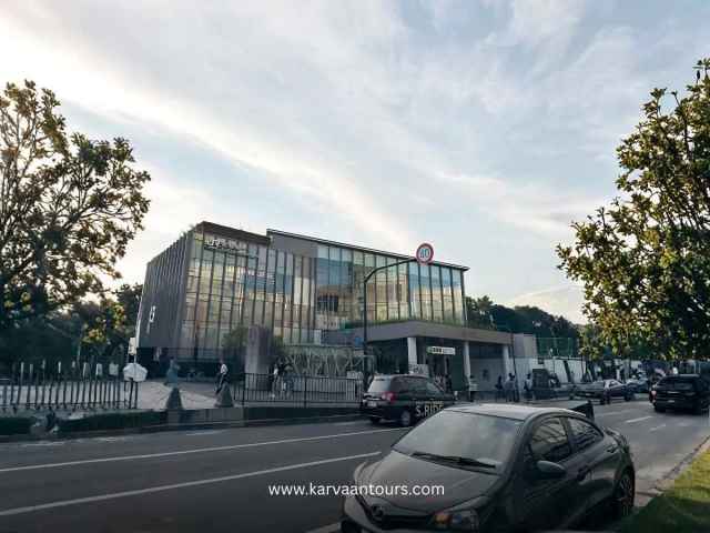 An exterior street-level view of the modern, glass-fronted Harajuku Station building in Tokyo during the late afternoon, with cars and taxis driving along the road in front.