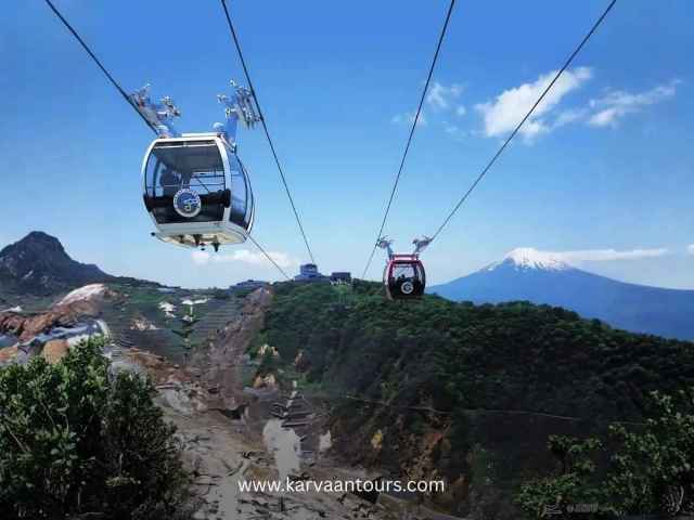 A gondola of the Hakone Ropeway traveling over the rugged, volcanic terrain of Owakudani, with Mount Fuji standing prominently in the distant horizon.