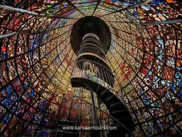 The iconic stained-glass tower at the Hakone Open-Air Museum, featuring a spiral staircase inside illuminated by vibrant, multi-colored light filtering through the intricate glass panels.