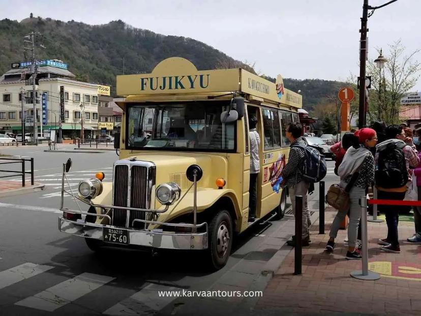 A vintage-style, pale yellow "Fujikyu" sightseeing bus waiting at a bus stop with a line of passengers preparing to board; a mountain landscape is visible in the background.