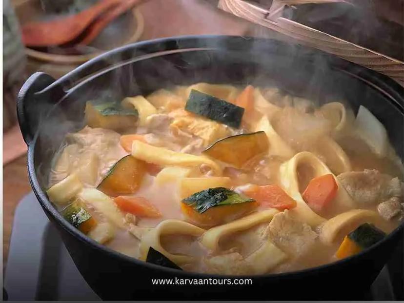 A close-up, high-angle shot of a steaming black pot of Hoto noodles—a local dish from the Fuji Five Lakes region—featuring thick noodles, pumpkin chunks, and carrots in a rich, savory broth.
