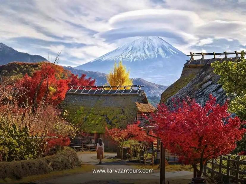 A woman walking along a path in a traditional Japanese village during autumn, surrounded by vibrant red and yellow foliage, with a magnificent snow-capped Mount Fuji under a distinctive lenticular cloud in the background.