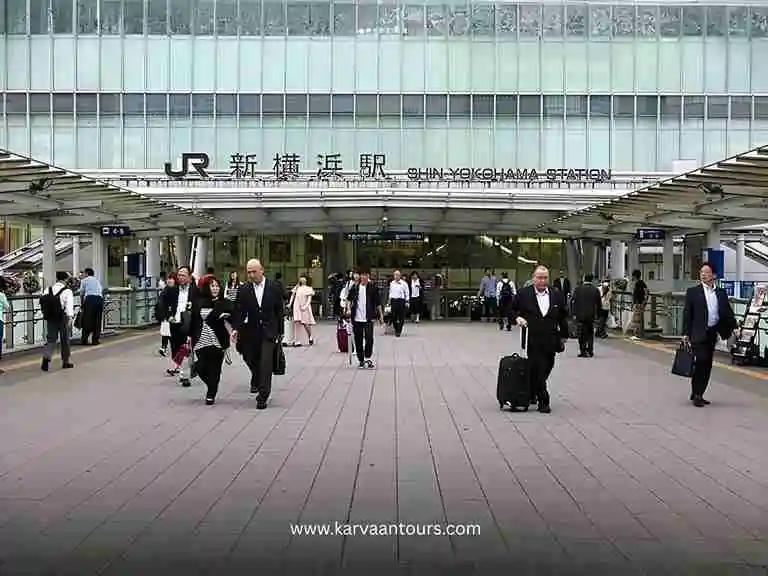 A busy daytime scene at the JR entrance of Shin-Yokohama Station, showing commuters and travelers walking through the bustling transit hub.