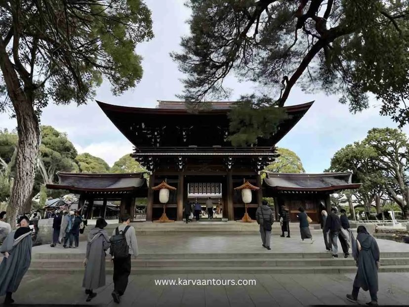 A view of the impressive main hall of Meiji Jingu Shrine in Tokyo, featuring traditional wooden architecture, large white hanging lanterns, and visitors walking through the courtyard under the shade of mature trees.
