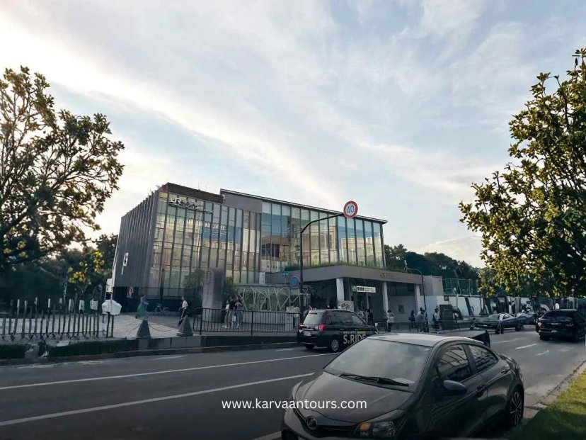 An exterior street-level view of the modern, glass-fronted Harajuku Station building in Tokyo during the late afternoon, with cars and taxis driving along the road in front.