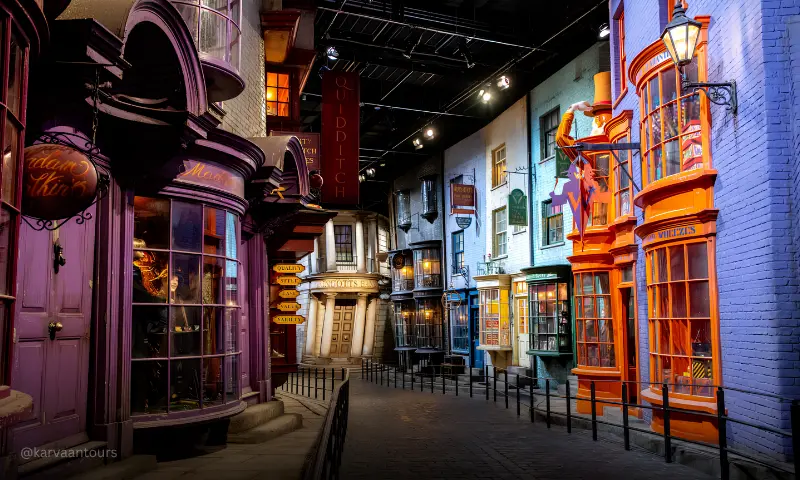A perspective shot looking down a cobbled wizarding street lined with colorful magical shops, including the bright purple storefront of Madam Malkin's and a vibrant orange Weasleys' Wizard Wheezes building beneath a dark indoor studio ceiling.