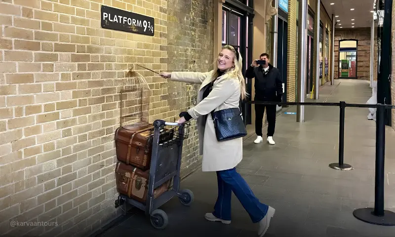 A smiling woman in a beige coat and blue jeans poses next to a brick wall, holding a magic wand and grabbing a luggage trolley loaded with vintage trunks and a birdcage that is halfway embedded into the wall under a "Platform 9 ¾" sign.