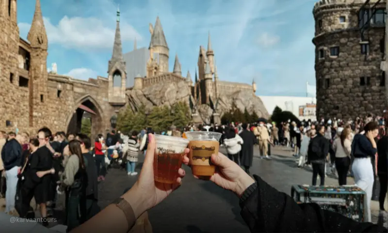 A first-person perspective shot of two hands clinking plastic cups filled with frothy Butterbeer. In the blurry background, a crowd of people walks around an outdoor courtyard with a large stone castle structure under a bright, partly cloudy sky.