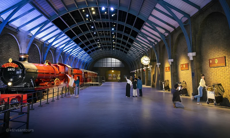 A wide view of a spacious indoor studio set replicating a train station platform. The crimson Hogwarts Express steam engine sits on the left, while visitors on the right take photos and pose with luggage carts disappearing into a brick wall under a high, arched blue ceiling.