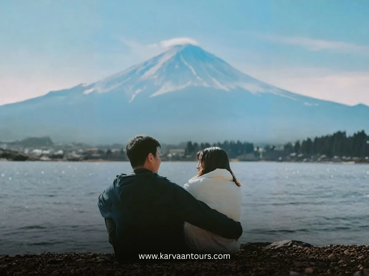A romantic, rear-view shot of a young couple sitting closely together on a pebbled shoreline, looking out over the calm waters of Lake Kawaguchiko toward a majestic, partially cloud-dusted Mount Fuji.