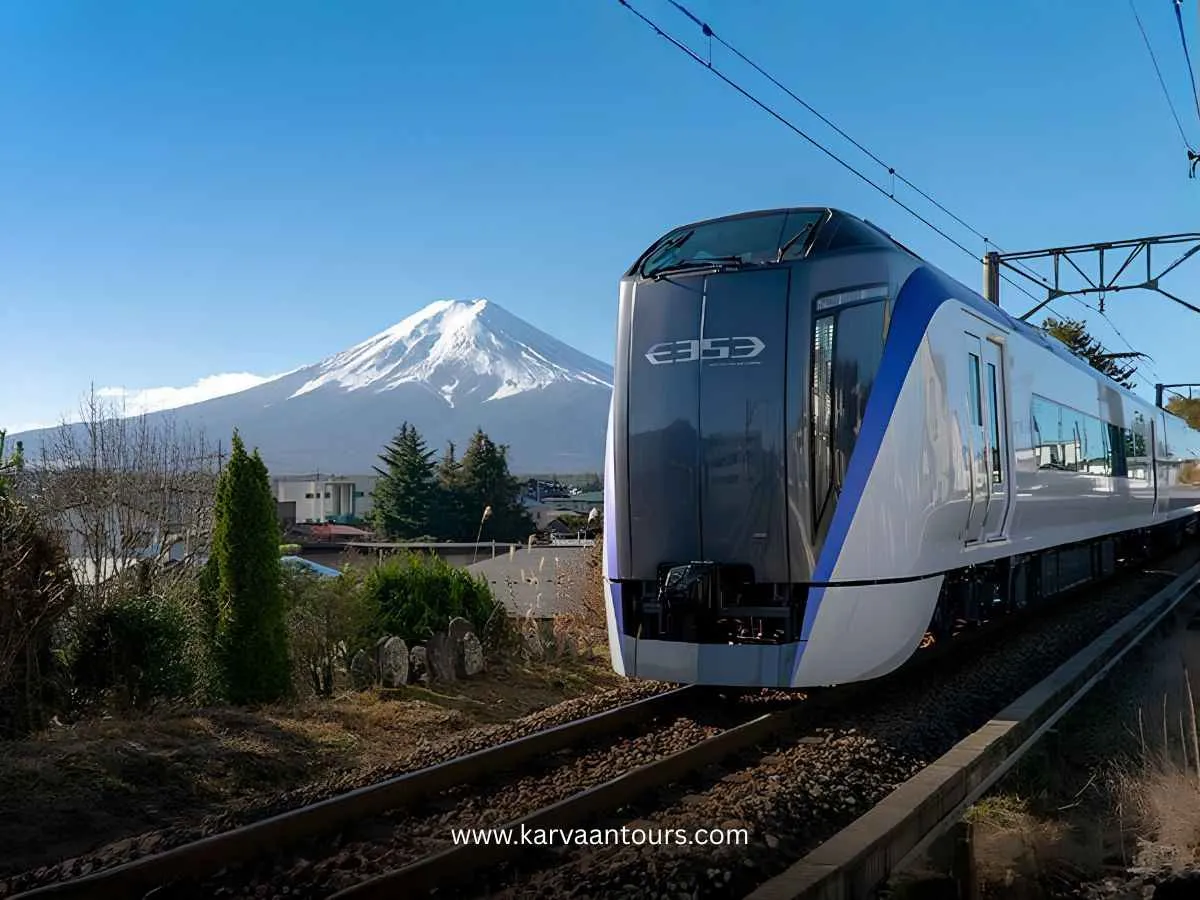 A modern, sleek white and blue passenger train, identified by the "E353" logo on its front, traveling along a railway track with a scenic, clear view of the snow-capped Mount Fuji in the background under a bright blue sky.