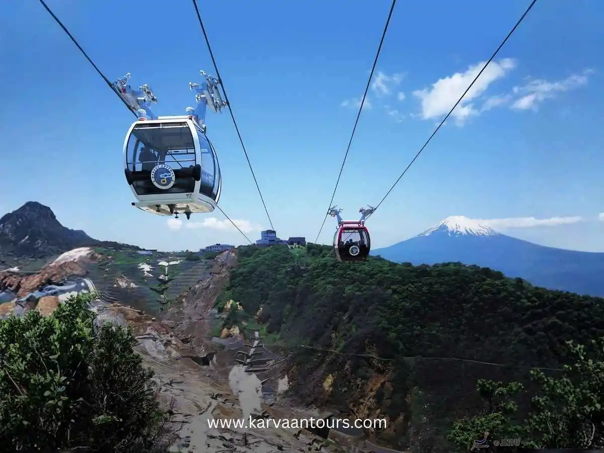 A gondola of the Hakone Ropeway traveling over the rugged, volcanic terrain of Owakudani, with Mount Fuji standing prominently in the distant horizon.