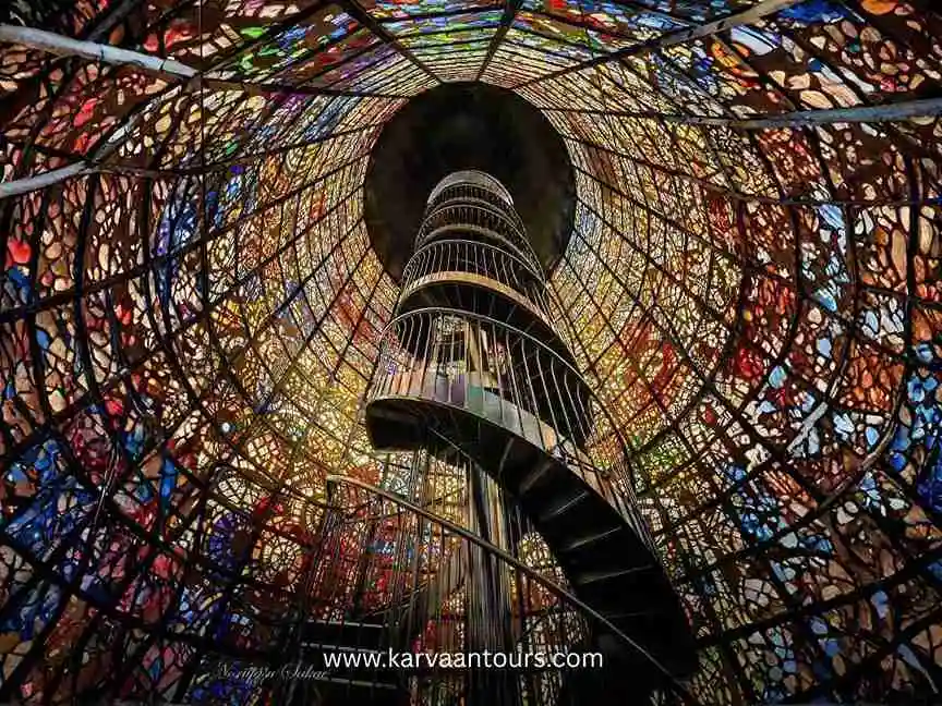 The iconic stained-glass tower at the Hakone Open-Air Museum, featuring a spiral staircase inside illuminated by vibrant, multi-colored light filtering through the intricate glass panels.
