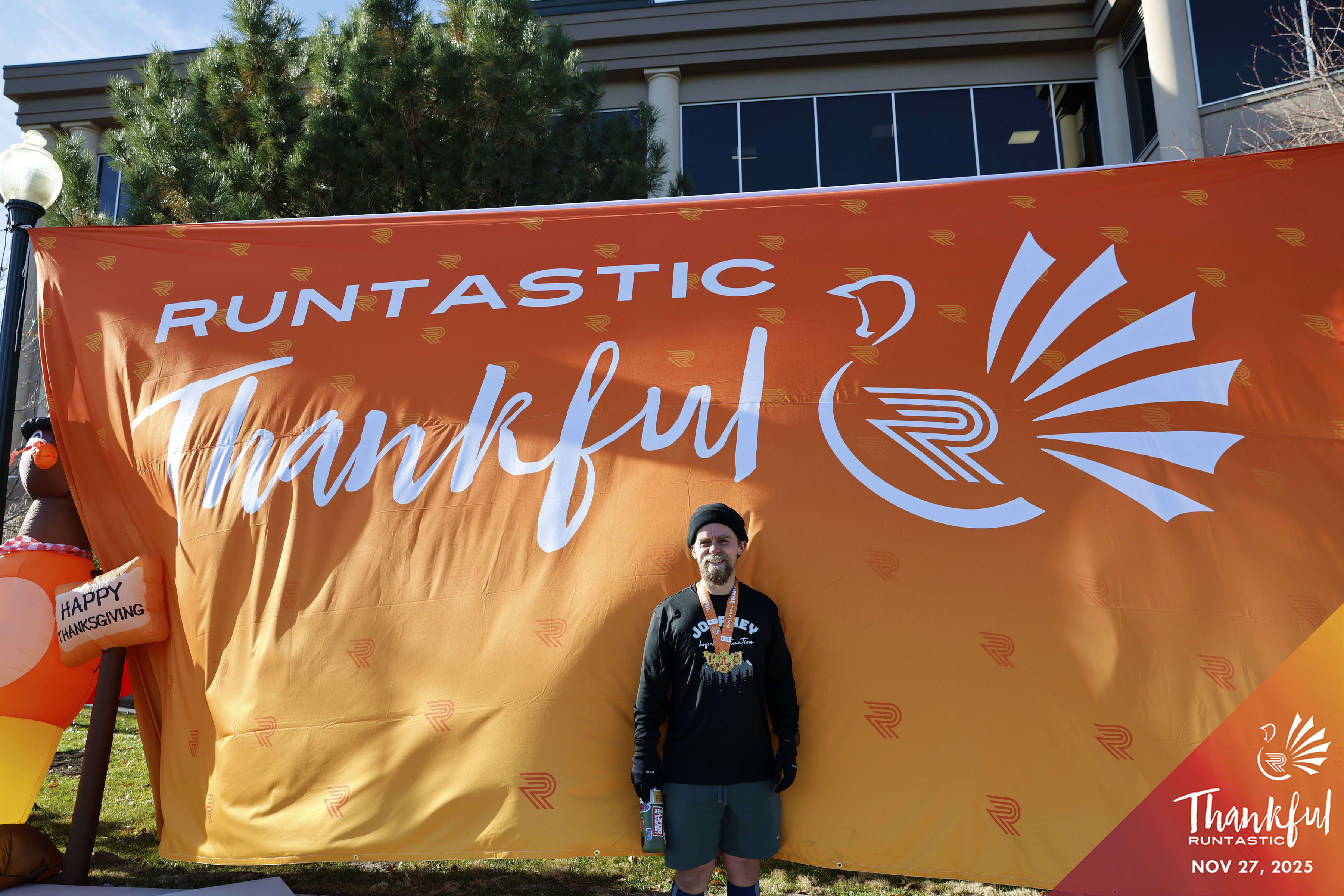 Andrew poses at a banner that says 'Runtastic Thankful'. There is an icon that has an R in the center and there is the drawing of a turkey surrounding the R.