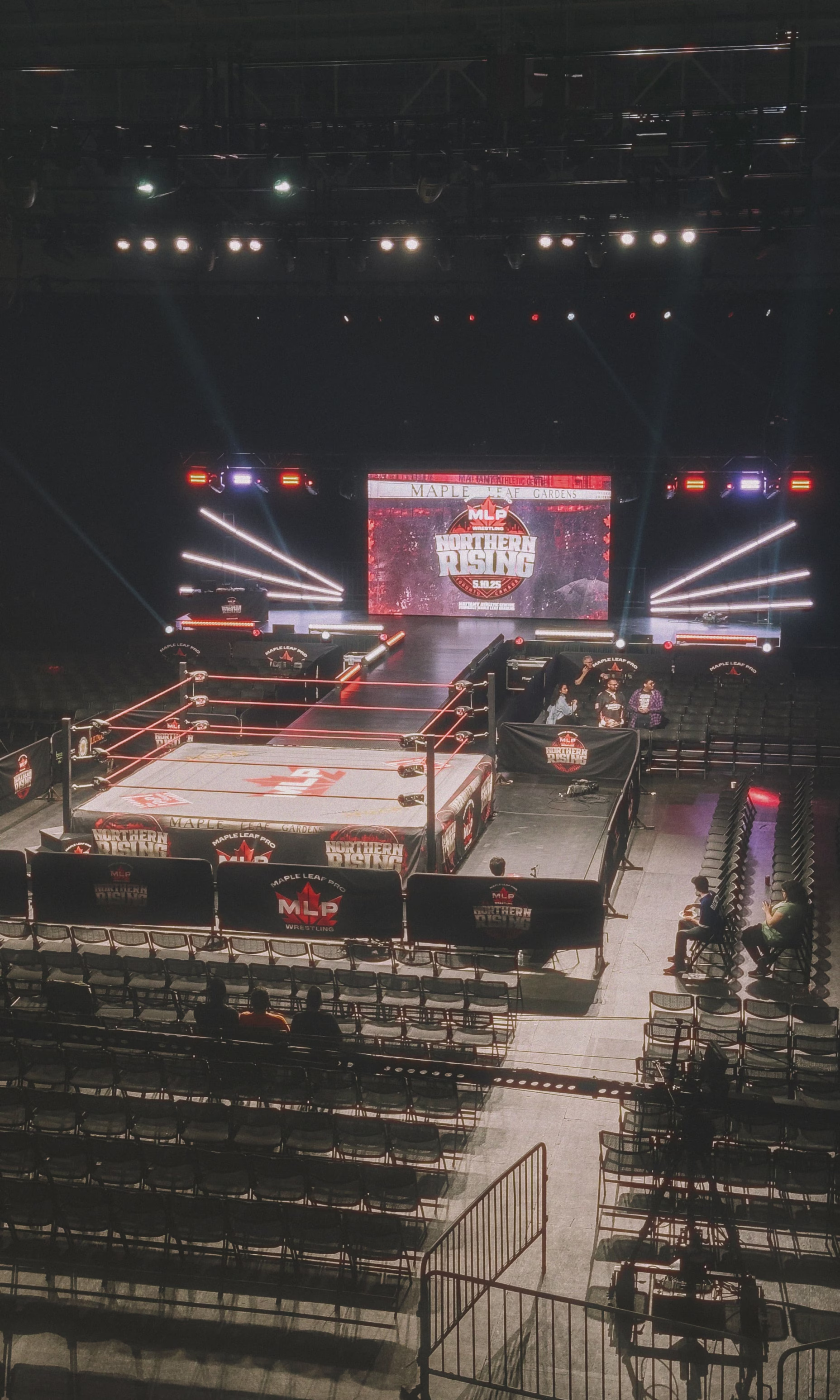 A photo of the interior of Maple Leaf Gardens, with the ring set up in the middle of the arena. The seats are mostly empty, with a few fans scattered around the front rows. The ring is lit up with bright lights, and the MLP logo is visible in the middle of the mat.