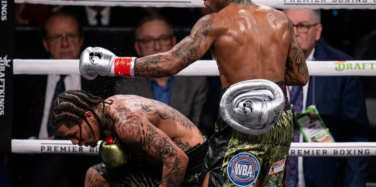 A close-up side profile of Gervonta Davis taking a knee on the canvas during a high-tempo round, looking up at the referee as the crowd in the background reacts wildly.