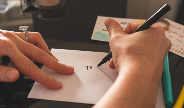 Close up of hands with a pen and wedding rings writing "To:" on an envelope