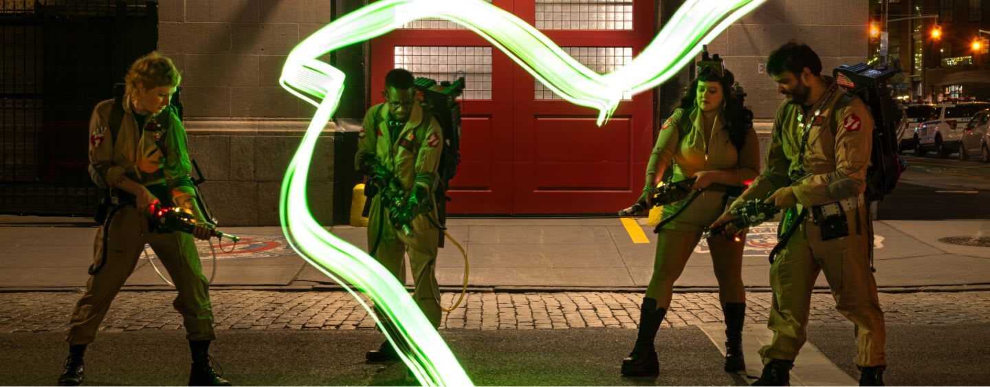 Four ghostbusters standing in front of Ghostbusters HQ, getting ready to fight a ghost coming out of a manhole.