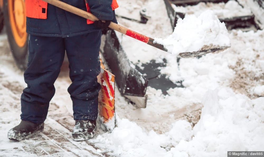Arbeiter schaufelt Schnee von Hand neben dem Räumschild eines Fahrzeugs