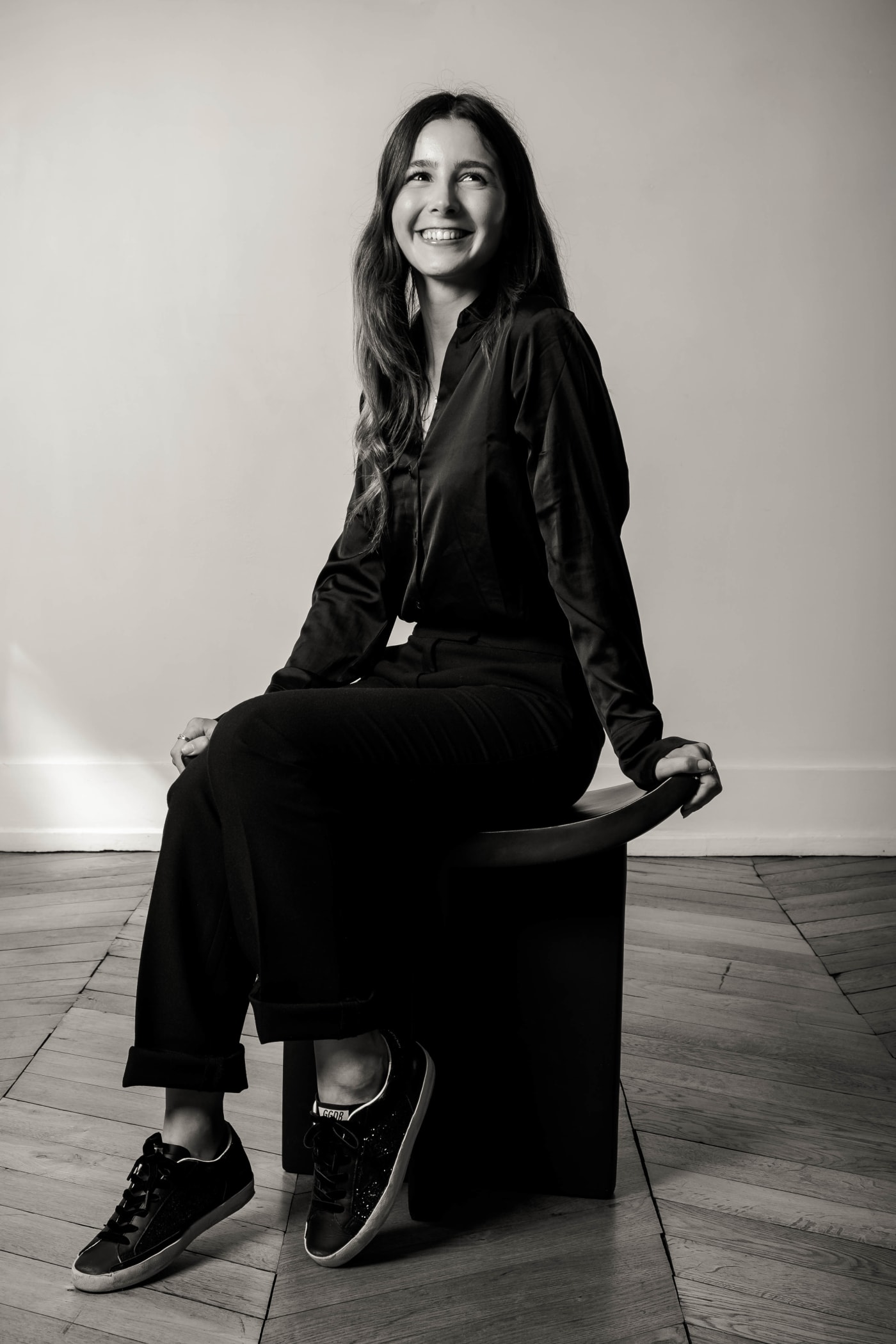 Designer souriante assise sur un tabouret noir, en tenue sombre, sur un parquet en chevrons