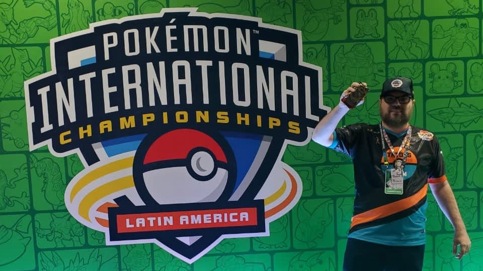 João Carlos Abitante, de boné e camisa do TATOYS TEAM, ergue o braço ao lado do painel do Pokémon International Championships Latin America