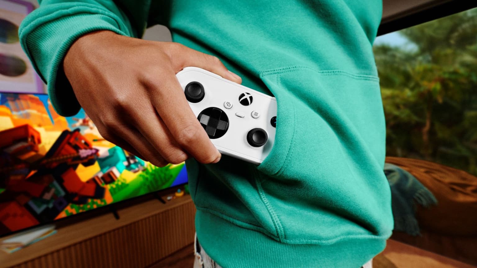 Mão tira o Controle Xbox Nano, branco, do bolso de um moletom verde, diante de uma TV ligada