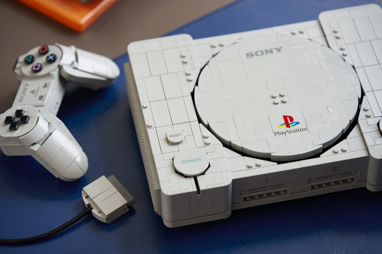 Réplica em LEGO do PlayStation original com o controle e o memory card conectados, sobre uma mesa azul