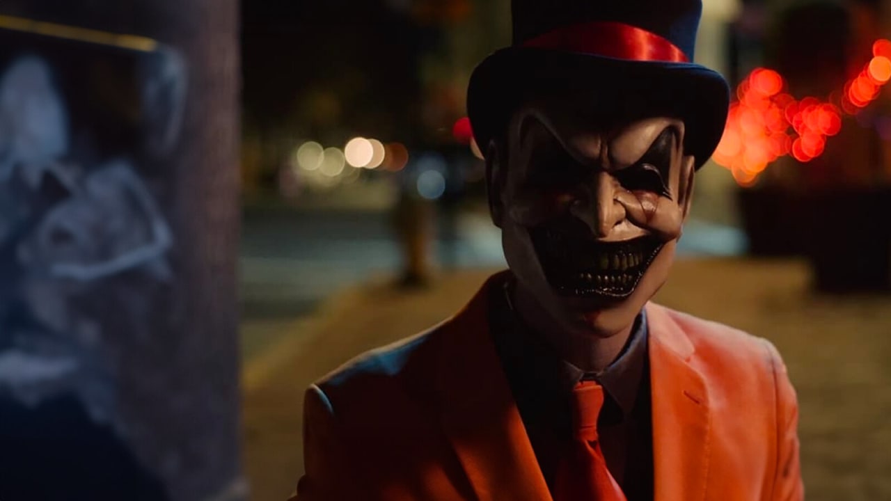 Figura de cartola, máscara de sorriso largo e terno laranja numa rua iluminada à noite