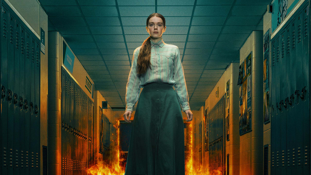 Carrie, de camisa listrada e saia longa, encara a câmera no corredor de uma escola enquanto o fogo sobe atrás dela entre os armários