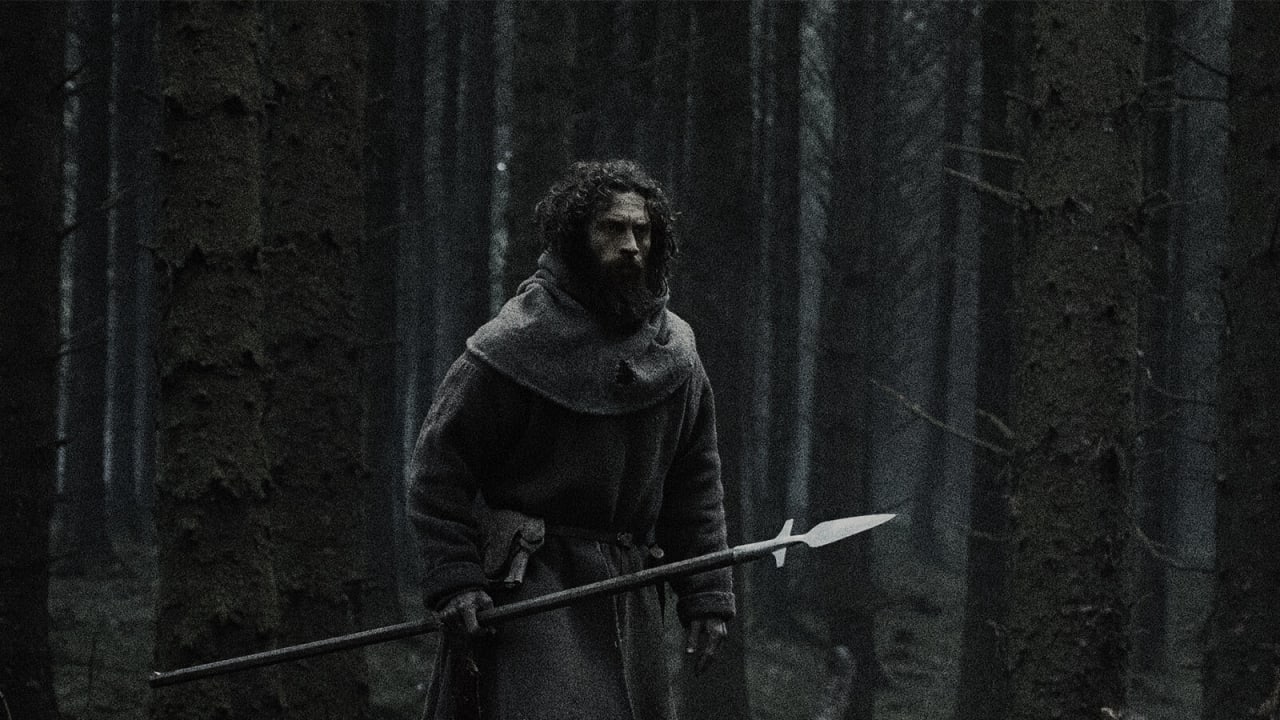 Aaron Taylor-Johnson, de capuz e barba, segura uma lança numa floresta escura de pinheiros