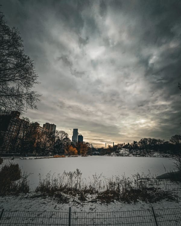 A snow-covered lake beneath dramatic clouds, with buildings along the far shore