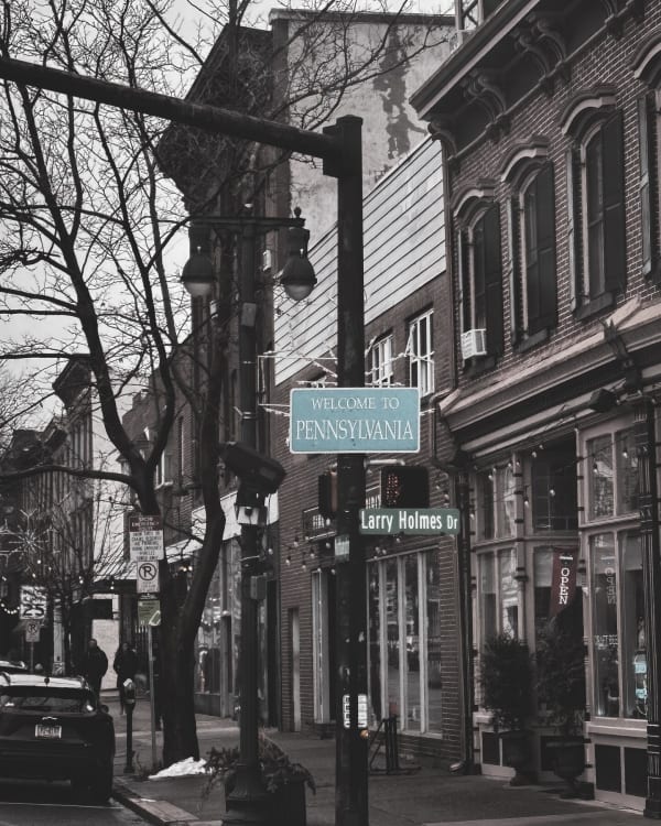 Black-and-white view of storefronts beside a Welcome to Pennsylvania sign