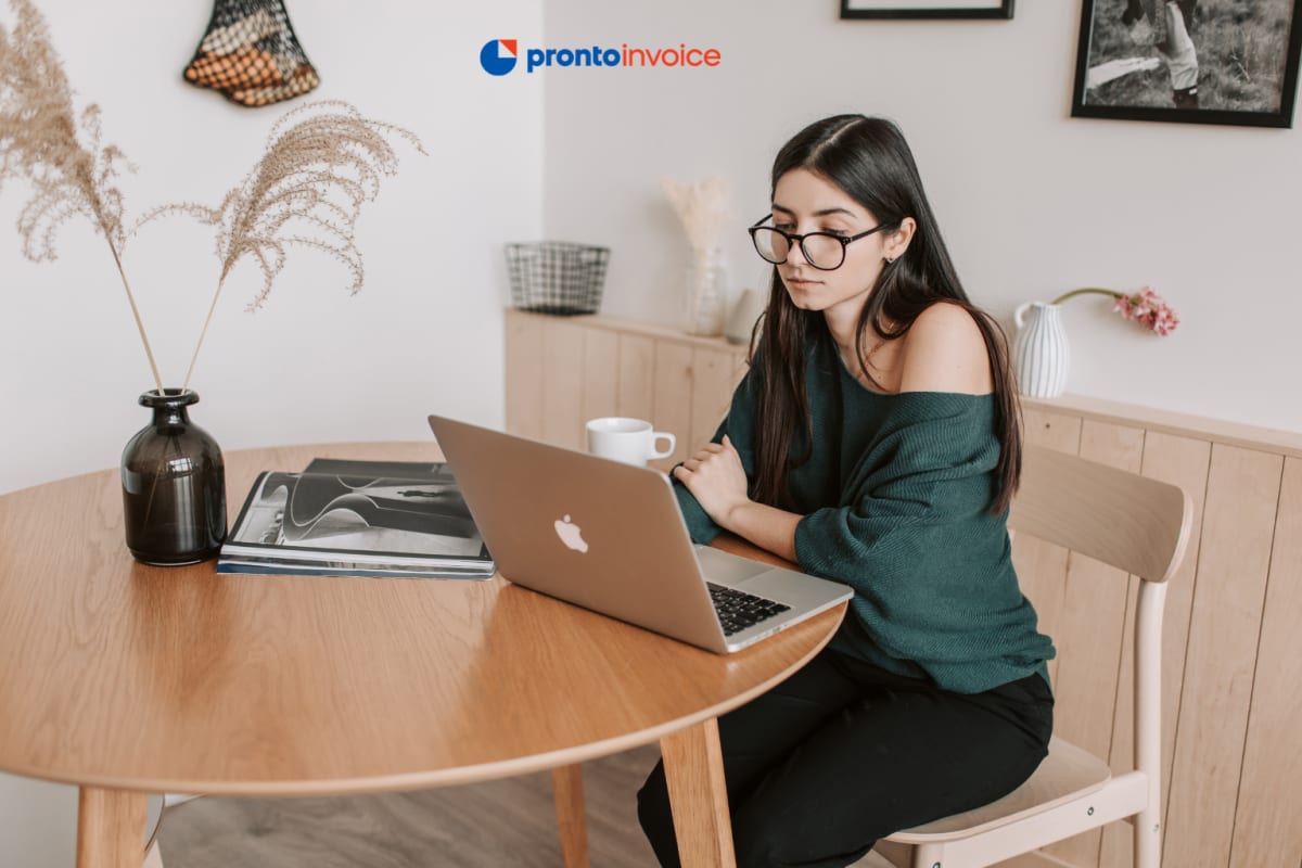 A young freelancer reviews their first invoice on a laptop at a home office desk in natural window light.
