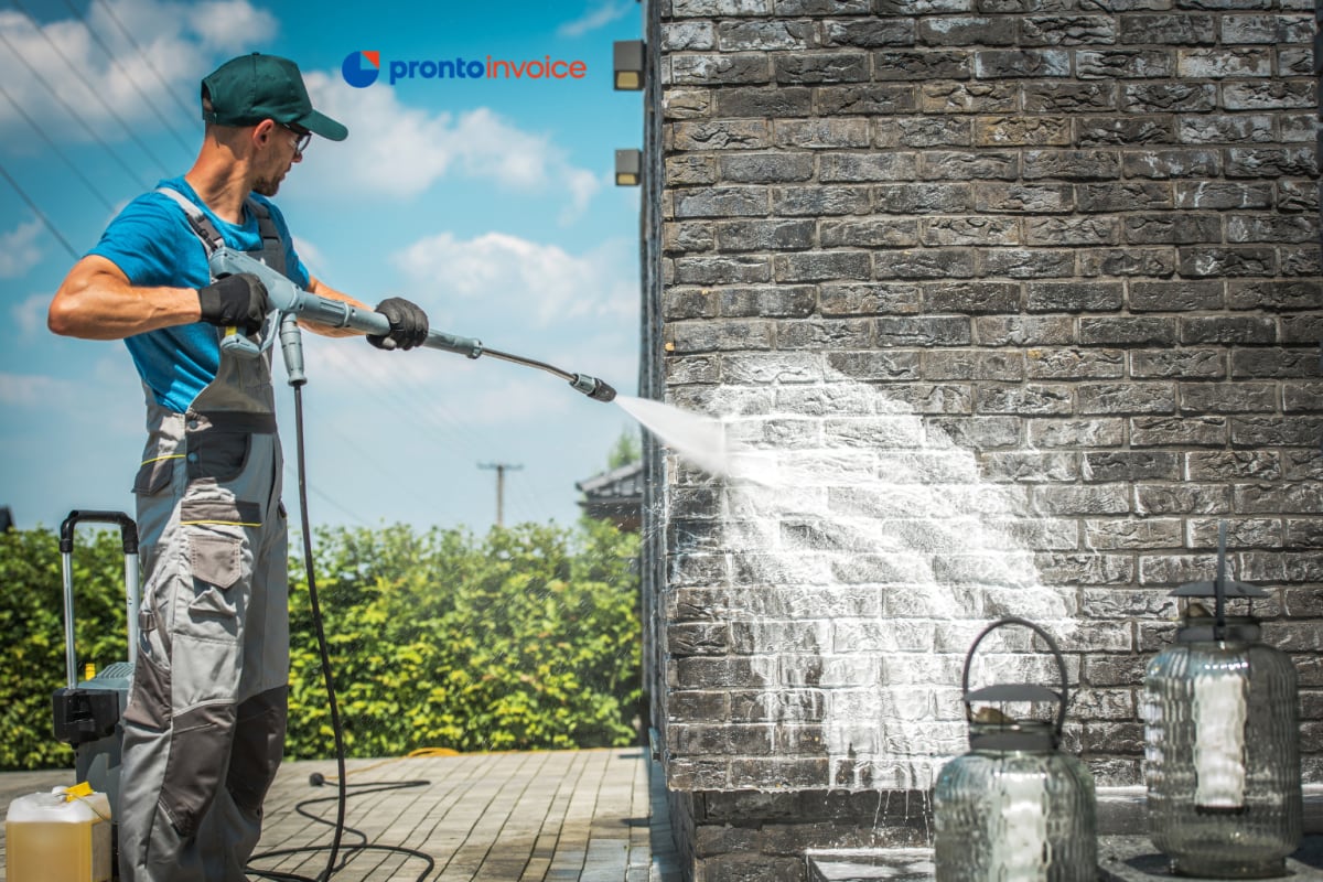 A pressure washing business owner in work clothes and safety goggles checks a job estimate on a phone beside a commercial pressure washer on a residential driveway in morning light.