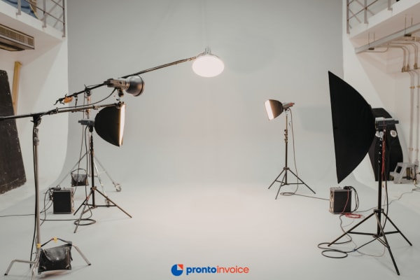 A photographer reviewing a session invoice on a smartphone at a studio desk with camera equipment and softboxes visible in the background.