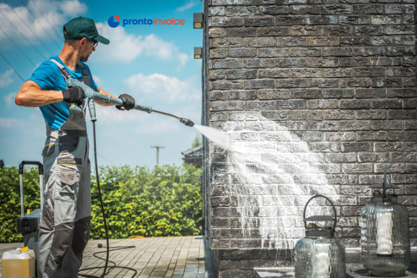 A pressure washing business owner in work clothes and safety goggles checks a job estimate on a phone beside a commercial pressure washer on a residential driveway in morning light.