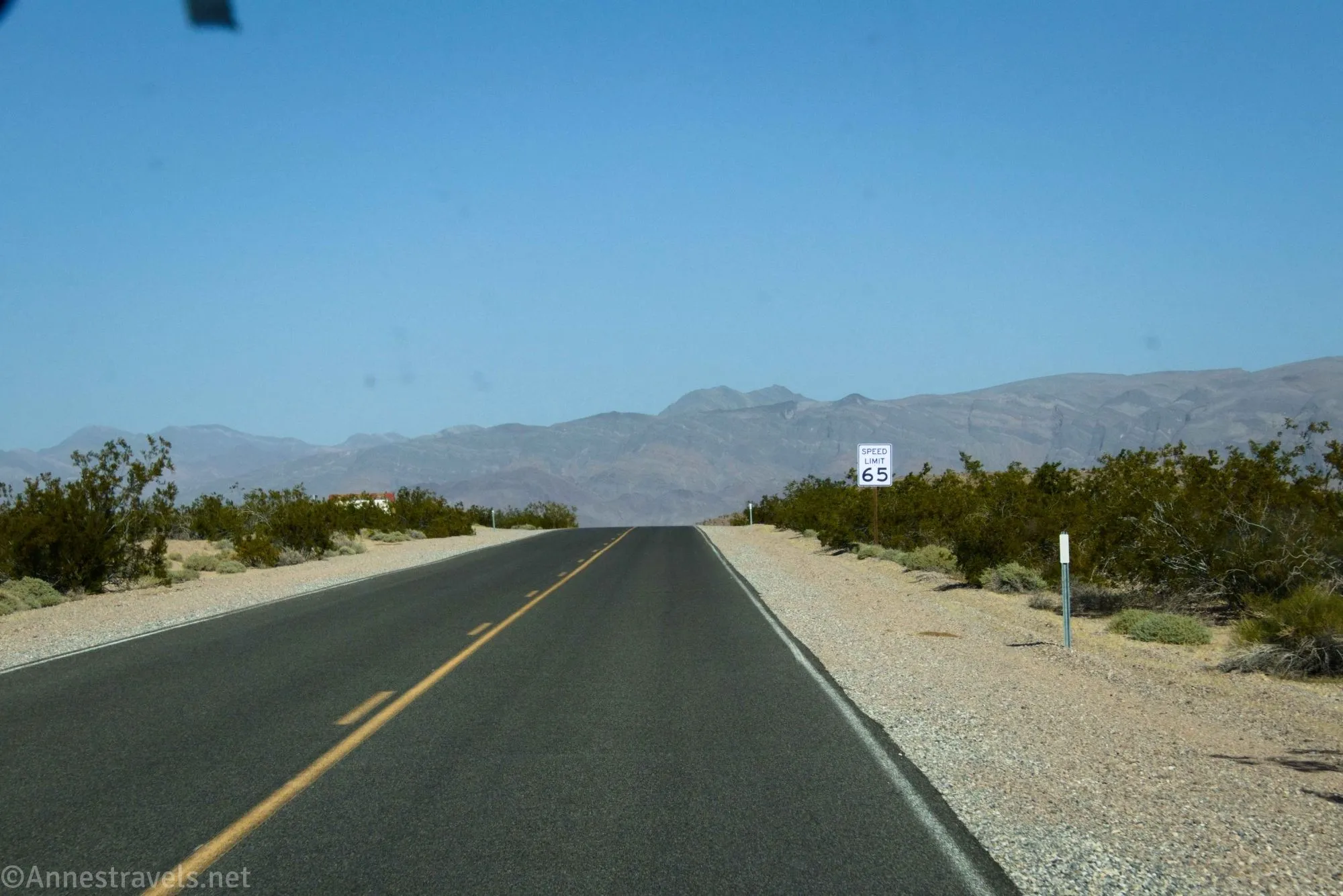 A paved road traveling straight through the desert between green greasebushes with a 65mph speed limit sign