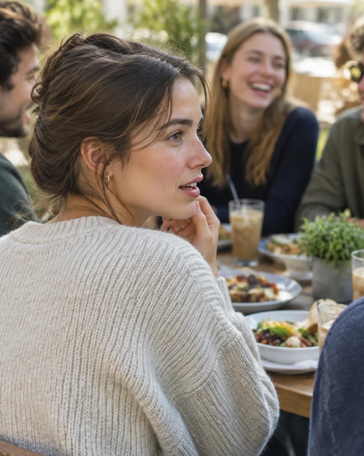 Young woman pausing mid-sentence at a cafe table while friends laugh behind her
