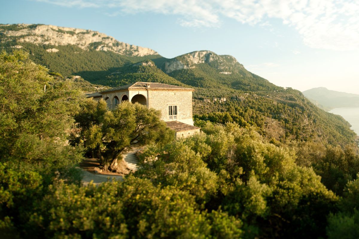 Sonnenbeschienene Finca umgeben von mallorquinischer Natur und Bergen im Hintergrund.