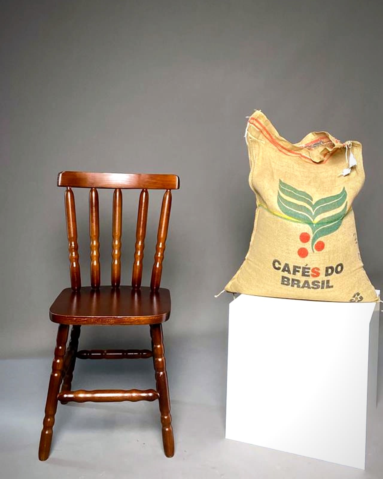 Saca de café verde apoiada num cubo branco, ao lado de uma cadeira de madeira