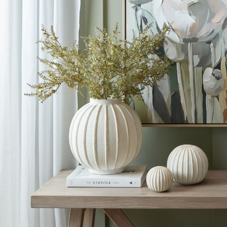 A cream-coloured matte ceramic globe vase with a ribbed texture displayed on a minimalist wooden side table.