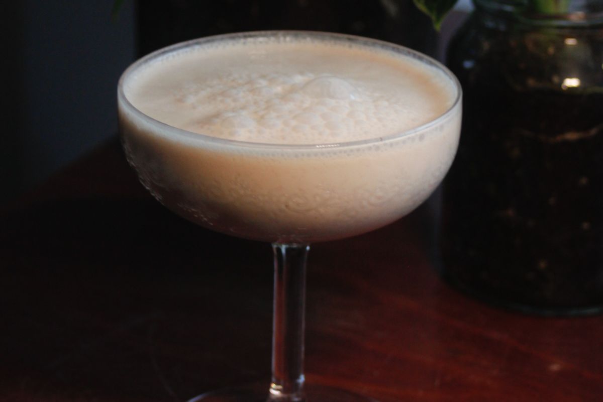 Ein Cocktailglas steht auf einem Holztisch und ist mit einem schaumigen, cremigen Getränk gefüllt. Im Hintergrund sind unscharf eine Pflanze und ein weiteres Glas zu sehen. Das Getränk wirkt frisch und einladend, perfekt für einen entspannten Abend.