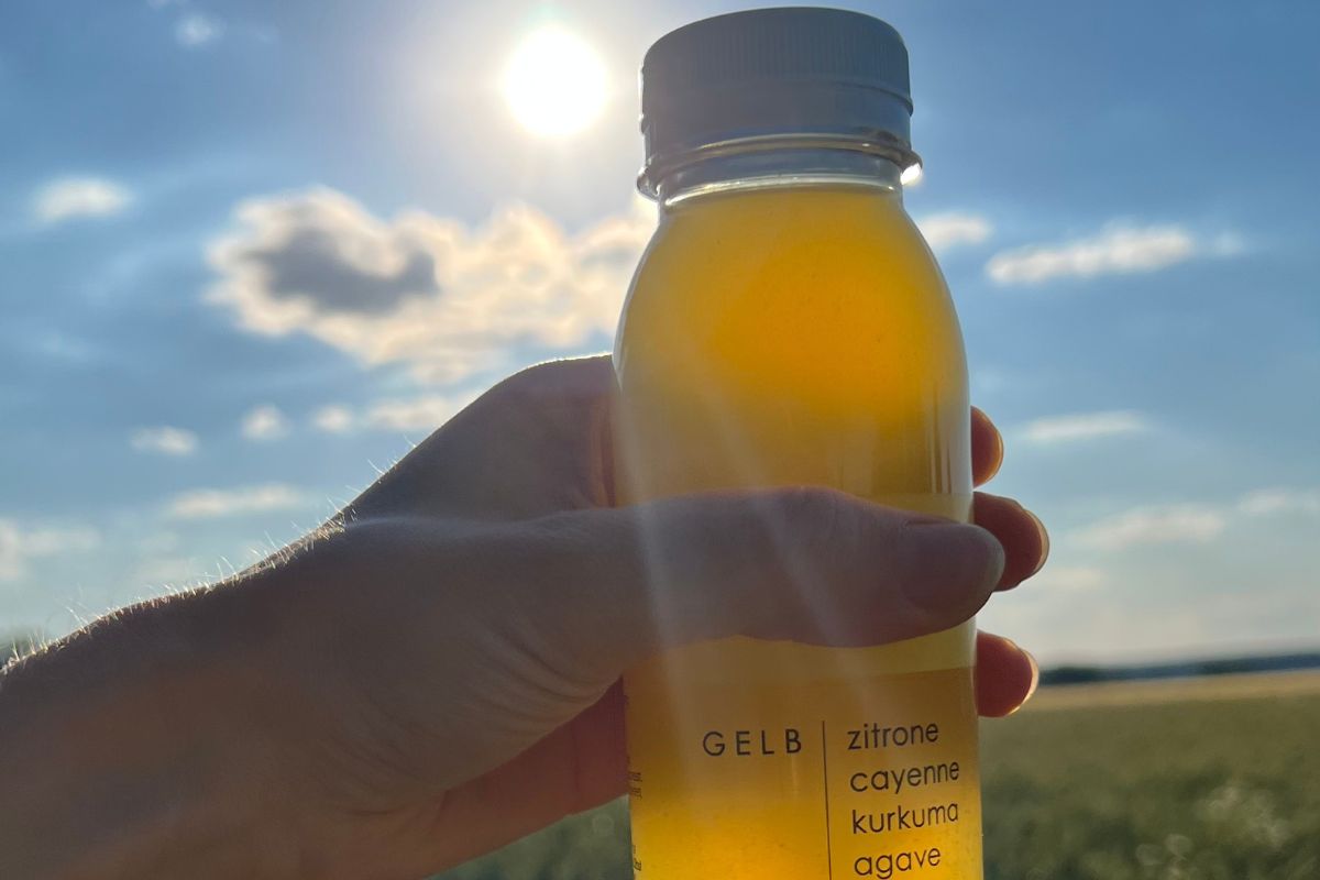 Eine Hand hält eine Flasche mit gelbem Getränk gegen das Sonnenlicht. Auf der Flasche steht "Zitrone, Cayenne, Kurkuma, Agave". Im Hintergrund ein strahlend blauer Himmel mit wenigen Wolken und die Sonne, die hell scheint.